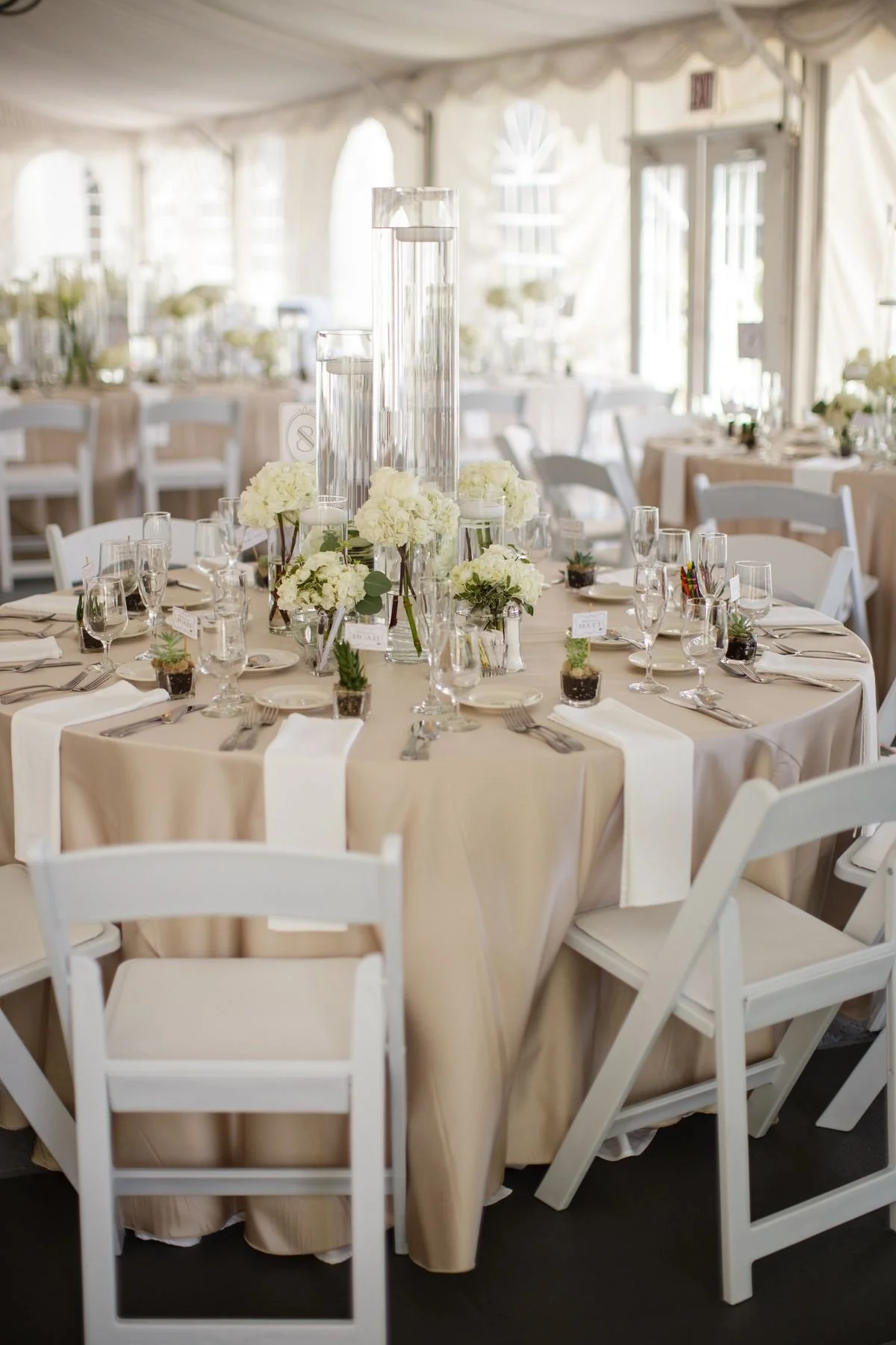 Elegant wedding reception table with a beige tablecloth, white folding chairs, and a centerpiece of white flowers in glass vases surrounded by tall glass cylinders filled with water, set in a white tent with natural light.