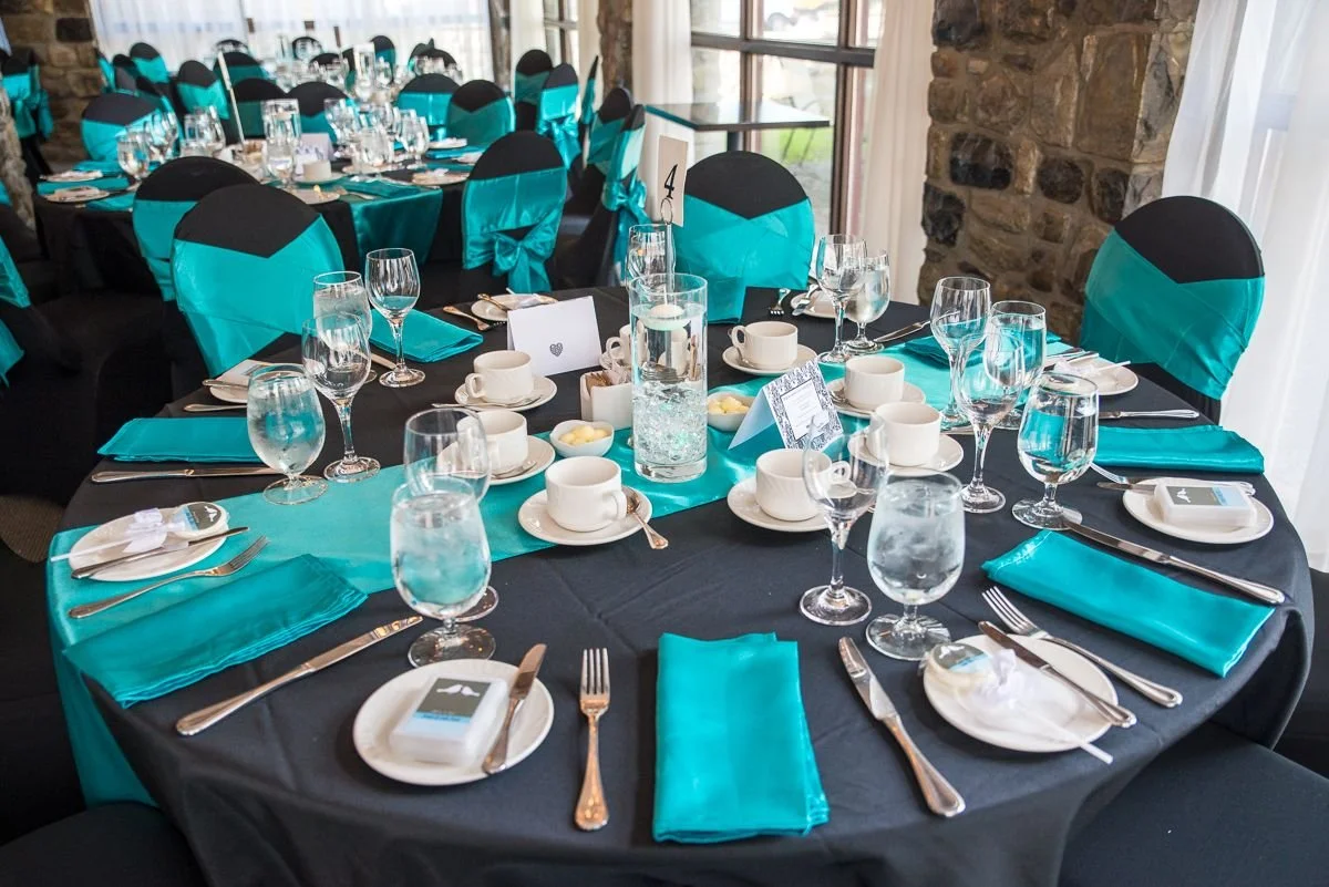 A round banquet table set for an event, covered with a black tablecloth, with black and turquoise chair covers. The table has turquoise and white accents, including napkins, chair sashes, and a table runner. There are multiple glasses, cups, plates, and silverware arranged around the table, along with a centerpiece consisting of a tall glass with water and floating candles, small bowls with butter, and table number card.
