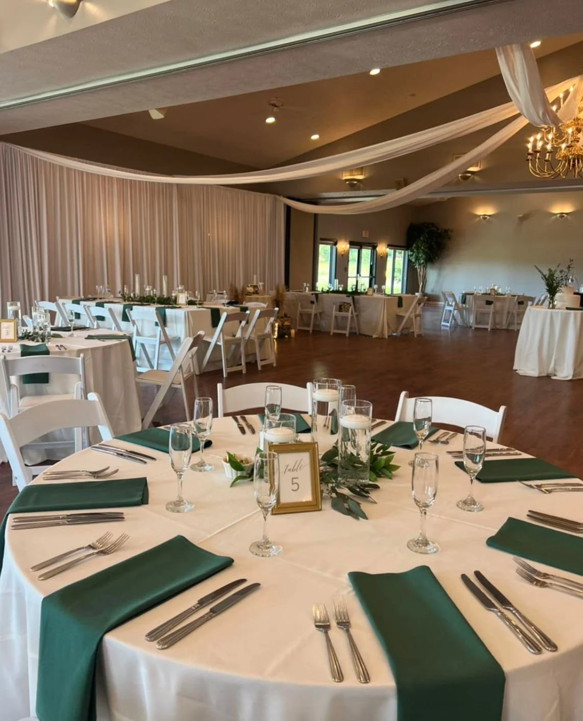 Wedding reception hall with round table set with green napkins, glassware, and silverware, and a table card labeled 'Table 5'. The hall has additional tables, white chairs, draped ceiling fabric, chandelier, and wall sconces.