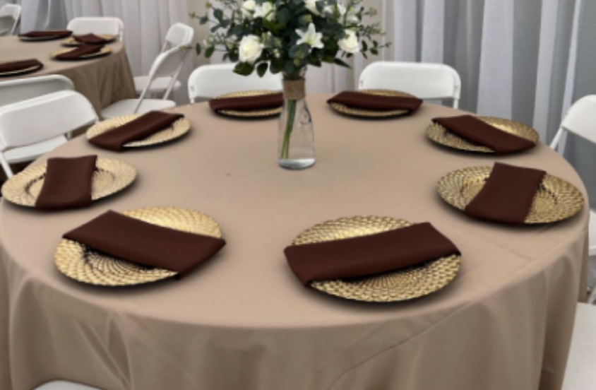 Round table set for a formal event with a beige tablecloth, gold and black woven placemats, brown napkins, and a central floral arrangement of white flowers and green foliage.