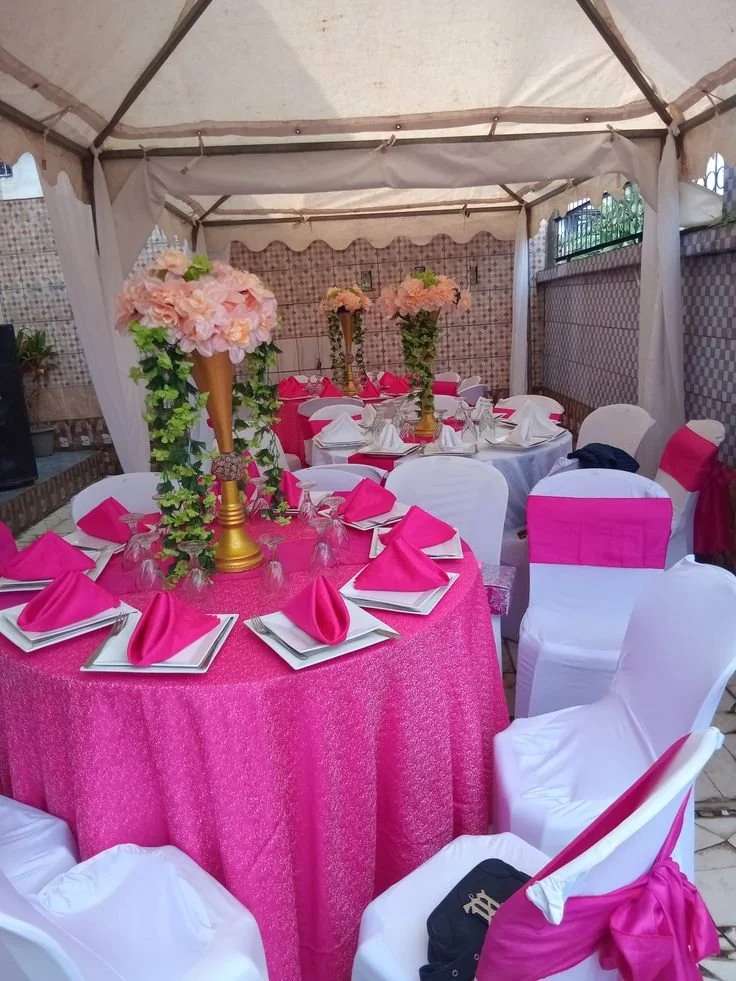 Decorated event tent with round tables covered in pink tablecloths and white chair covers with pink sashes, set for a celebration with pink floral centerpieces.