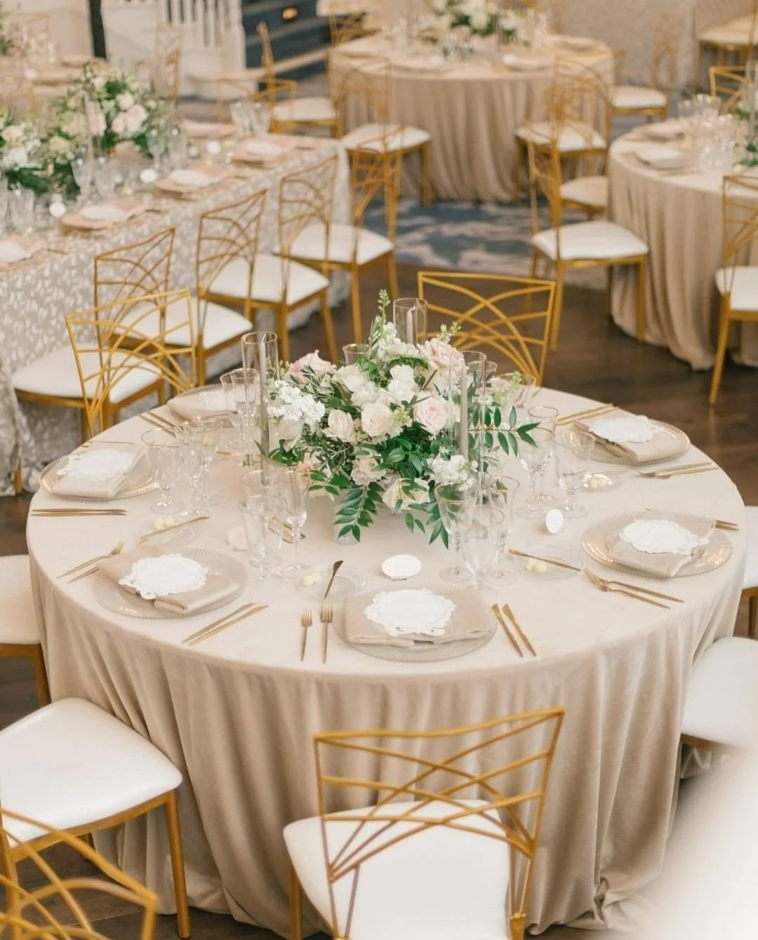 Elegant wedding reception table with white tablecloth, gold chairs, and floral centerpiece of white and pink roses and greenery, set with glassware, gold cutlery, and white plates.