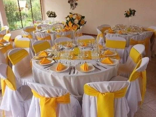 Round banquet table set with white tablecloth and yellow napkins, arranged with plates, silverware, and glasses, with yellow chair sashes, in a decorated event space with floral centerpieces.