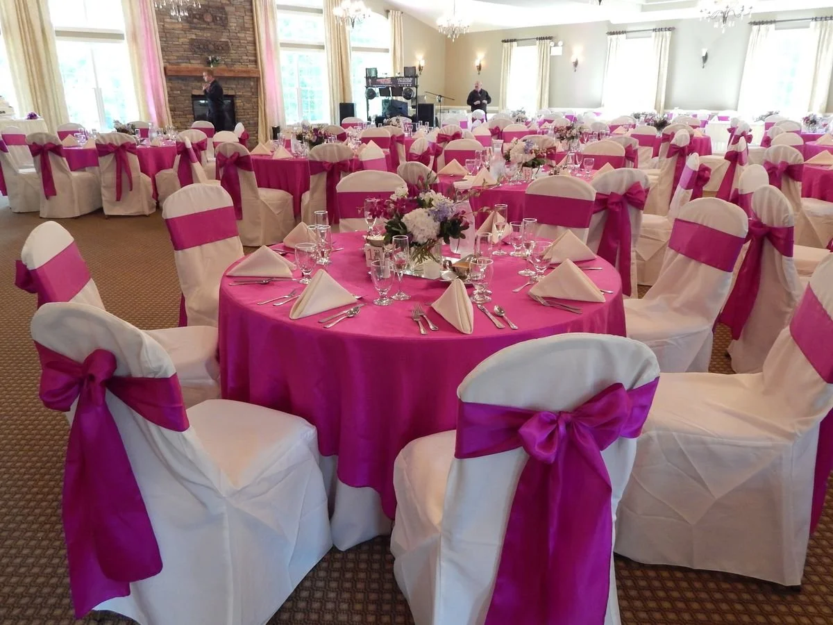 Decorated banquet hall with round tables covered with pink tablecloths and white chairs with pink sashes, set with glasses, cutlery, and napkins, and floral centerpieces.