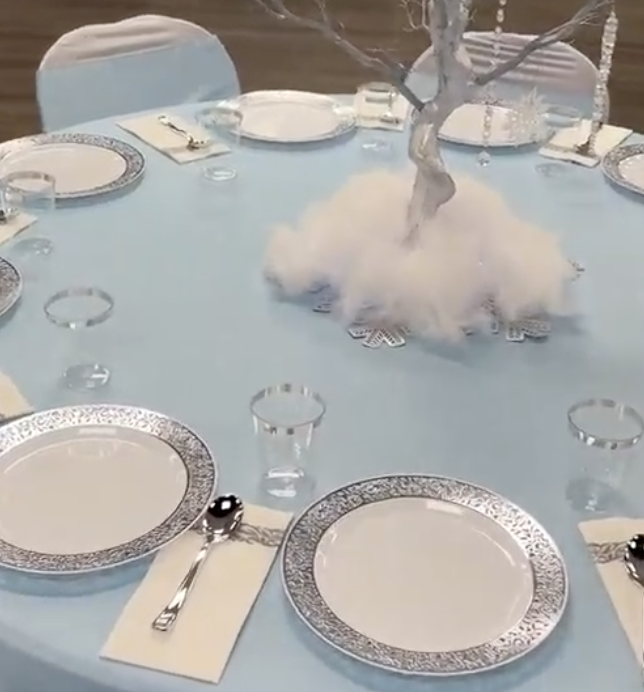 Elegant table setup with a light blue tablecloth, ornate silver-rimmed plates, clear glassware, and a centerpiece featuring a decorated white tree with hanging beads, surrounded by a fluffy white cushion.