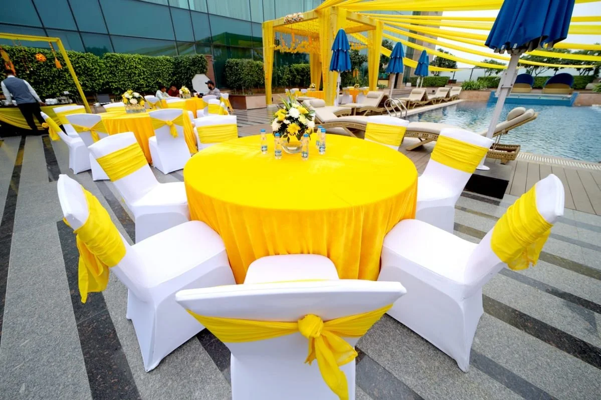 Outdoor poolside party setup with round tables covered in yellow tablecloths and white chairs decorated with yellow sashes, a pool visible in the background, and blue umbrellas providing shade.
