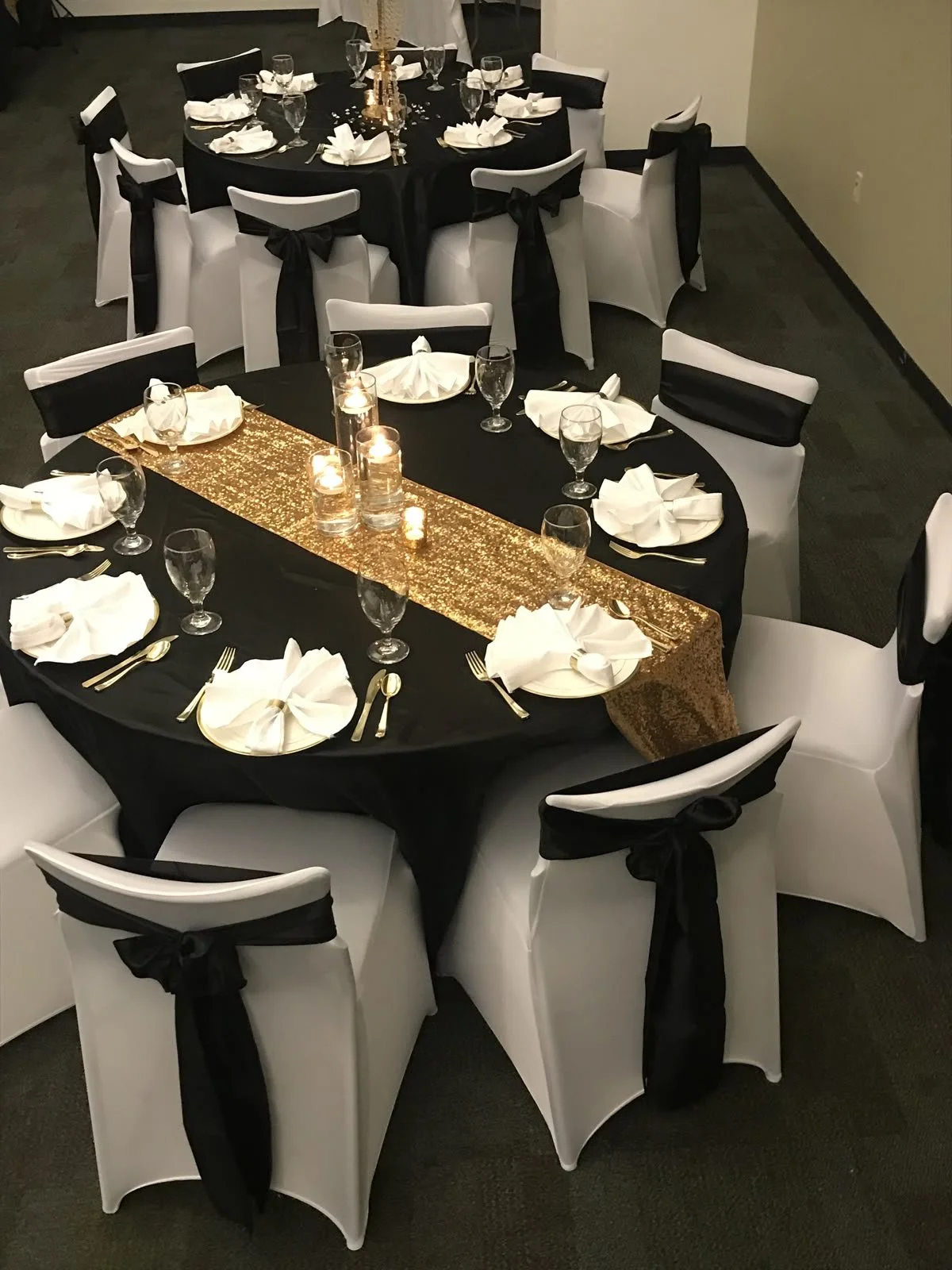 Round banquet tables decorated for a formal event, with black tablecloths, gold and black table runners, white chairs with black sashes, and candle centerpieces.