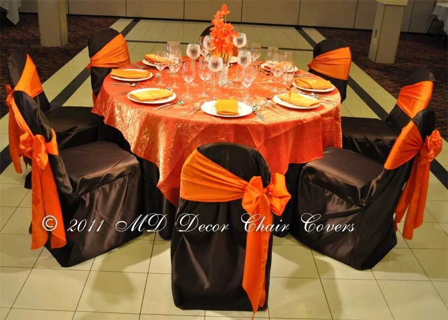 Round banquet table with orange tablecloth, set with glassware, plates, and yellow napkins, surrounded by six black chairs with orange sash covers.