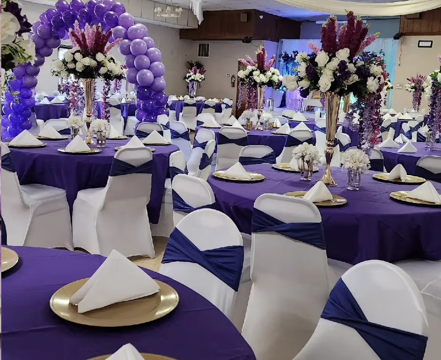 Elegant banquet hall decorated with purple and white theme, featuring round tables with purple tablecloths, white chairs with purple sashes, and tall floral centerpieces with white and purple flowers and balloons.