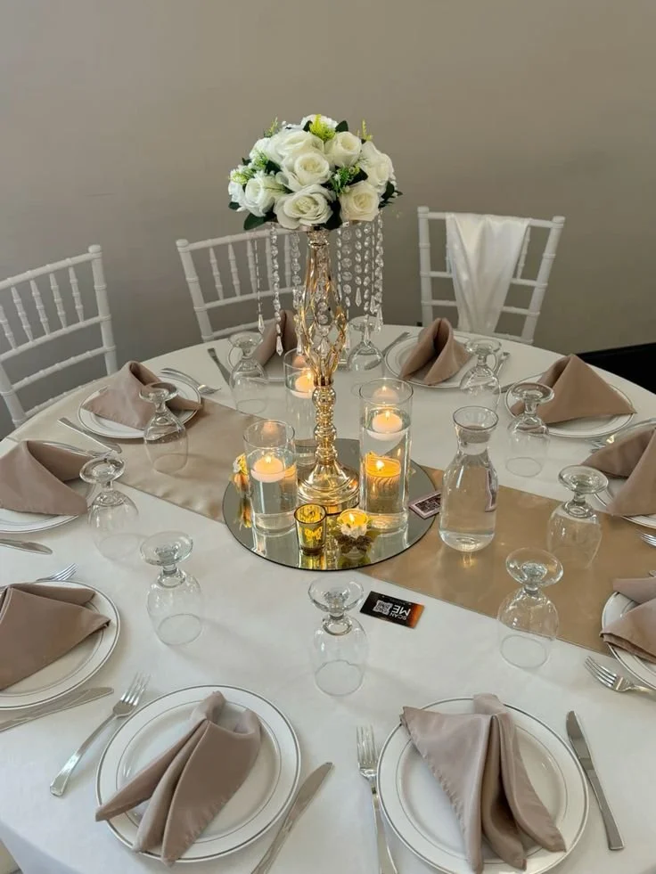 Formal dinner table setup with white tablecloth, beige napkins, clear wine glasses, water glasses, and a floral centerpiece with white roses and decorative hanging crystals, lit candles, and a small card.