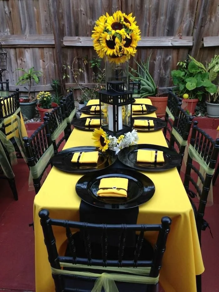 Outdoor dining table decorated for a celebration with yellow and black theme, featuring a sunflower centerpiece in a tall glass vase, yellow napkins, and black plates. The table is surrounded by black chairs with yellow ribbons, and the background includes potted plants and a wooden fence.