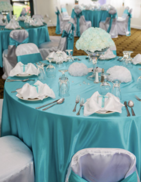 Wedding reception tables decorated with turquoise tablecloths, white napkins, glassware, and floral centerpieces of white flowers.