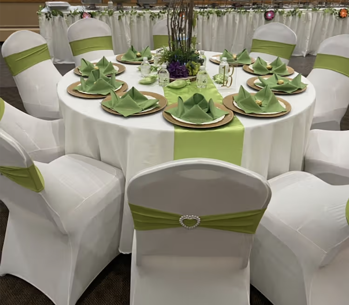 Round table decorated for a formal event with white tablecloth, green table runner, and green napkins folded in intricate design. White chairs with green sash adornments surround the table, which has a floral centerpiece with purple and green flowers.