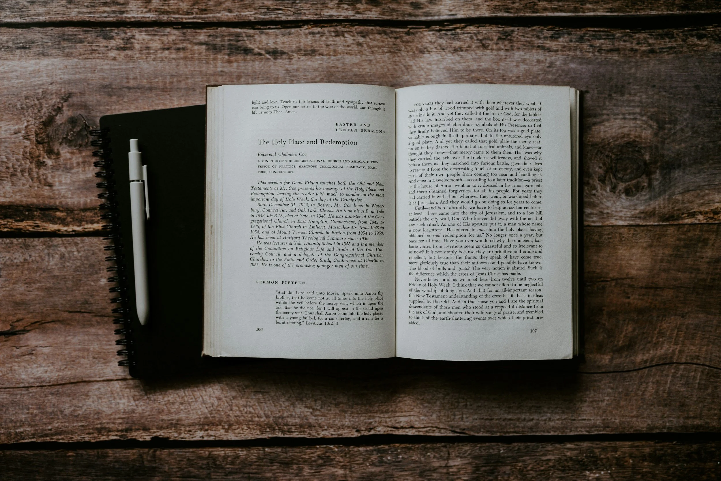 An open book on a wooden table next to a white pen.