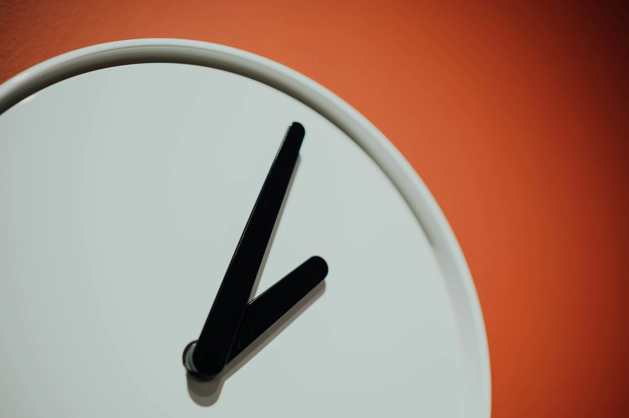 Close-up of a modern analog clock face with black clock hands on a white background, showing a time near 2:08, set against an orange background.