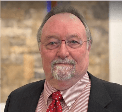 A middle-aged man with glasses, a beard, and a mustache, wearing a suit and red tie, standing against a blurred background of a brick wall.