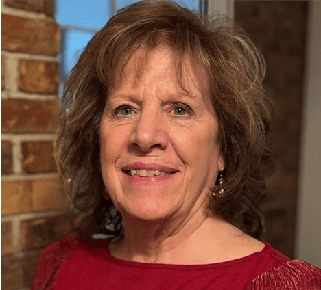 A middle-aged woman with light brown, curly hair and earrings, smiling, wearing a red top, standing indoors near a brick wall.