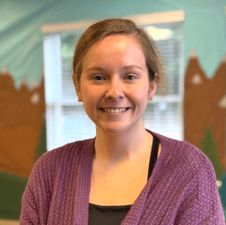 A woman with short light brown hair smiling, wearing a purple cardigan and a black top, standing in front of a colorful, mountain-themed mural with windows and trees.