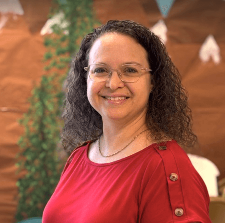 A woman with curly brown hair and glasses, wearing a red top with buttons on the shoulder, standing indoors with a wooden wall, green plant, and decorative bunting in the background.