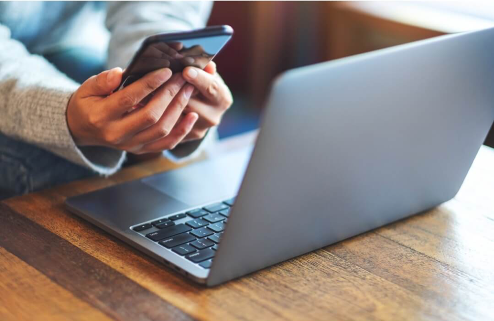 Person using a smartphone with a laptop on a wooden table.
