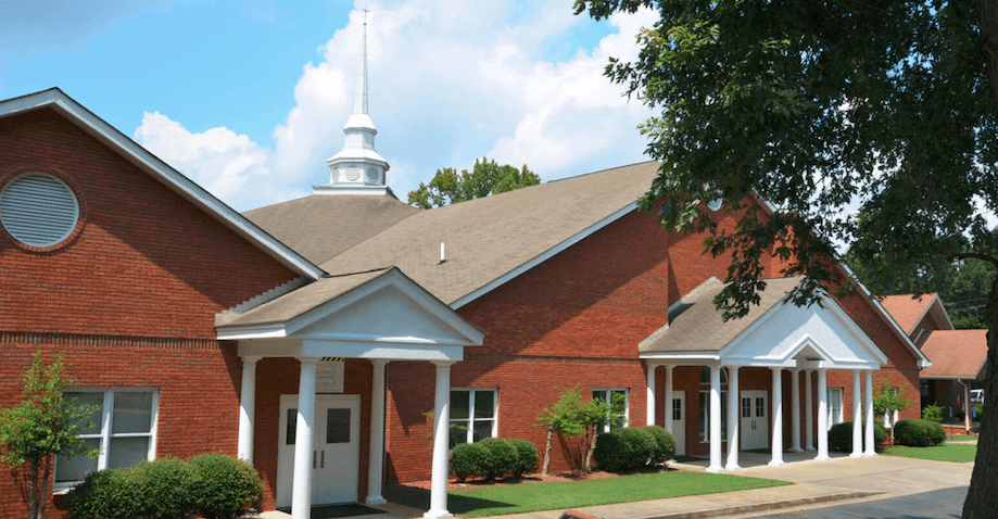 Red brick church building with white trim and columns, green bushes, and a large tree in the foreground under a partly cloudy sky.