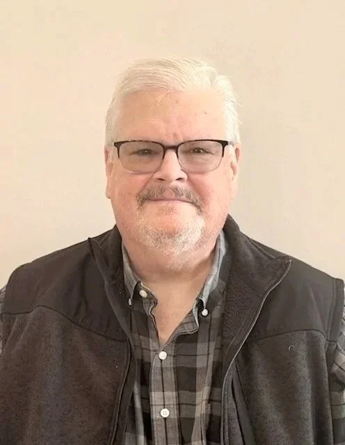 A man with glasses and white hair smiling, wearing a plaid shirt and black vest, standing against a plain beige wall.