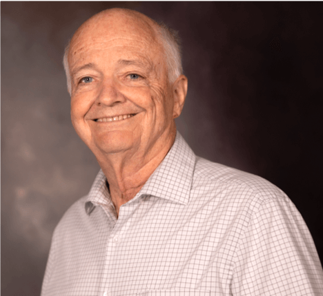 Older man with short white hair smiling, wearing a light-colored checkered shirt, against a dark background.