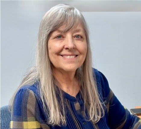 Smiling woman with long gray hair wearing a plaid shirt, sitting at a table.
