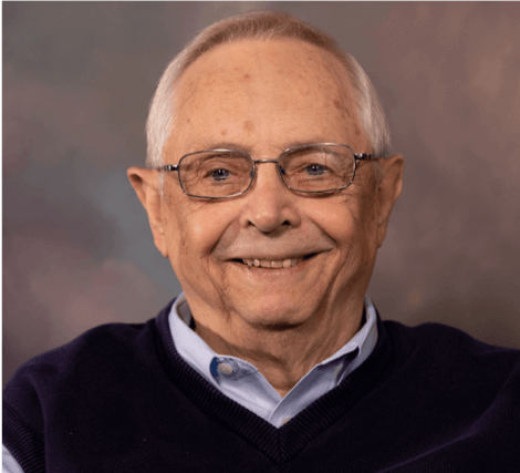 A smiling elderly man with glasses wearing a collared shirt and sweater, posed against a neutral background.