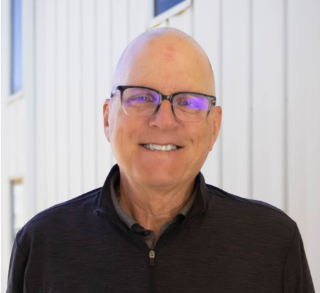 A man with glasses, wearing a dark blazer and a red plaid shirt, standing indoors near a window with natural light, smiling.