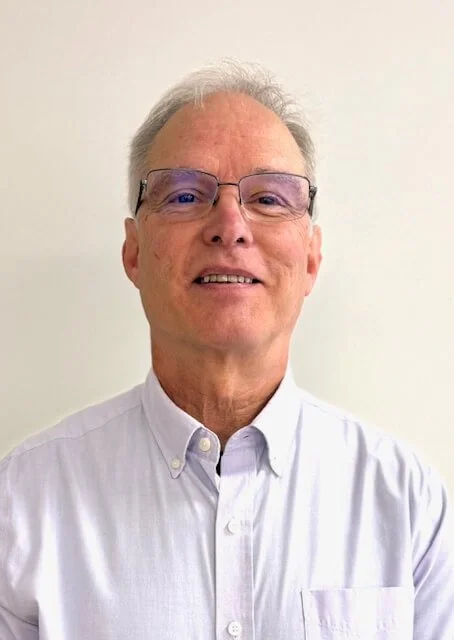 An older man with gray hair, glasses, and a white collared shirt standing against a plain white wall.