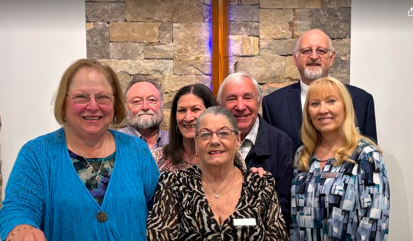 Group of seven people, four women and three men, standing together indoors, smiling. They are dressed in business casual attire, with a cross on a wall behind them.