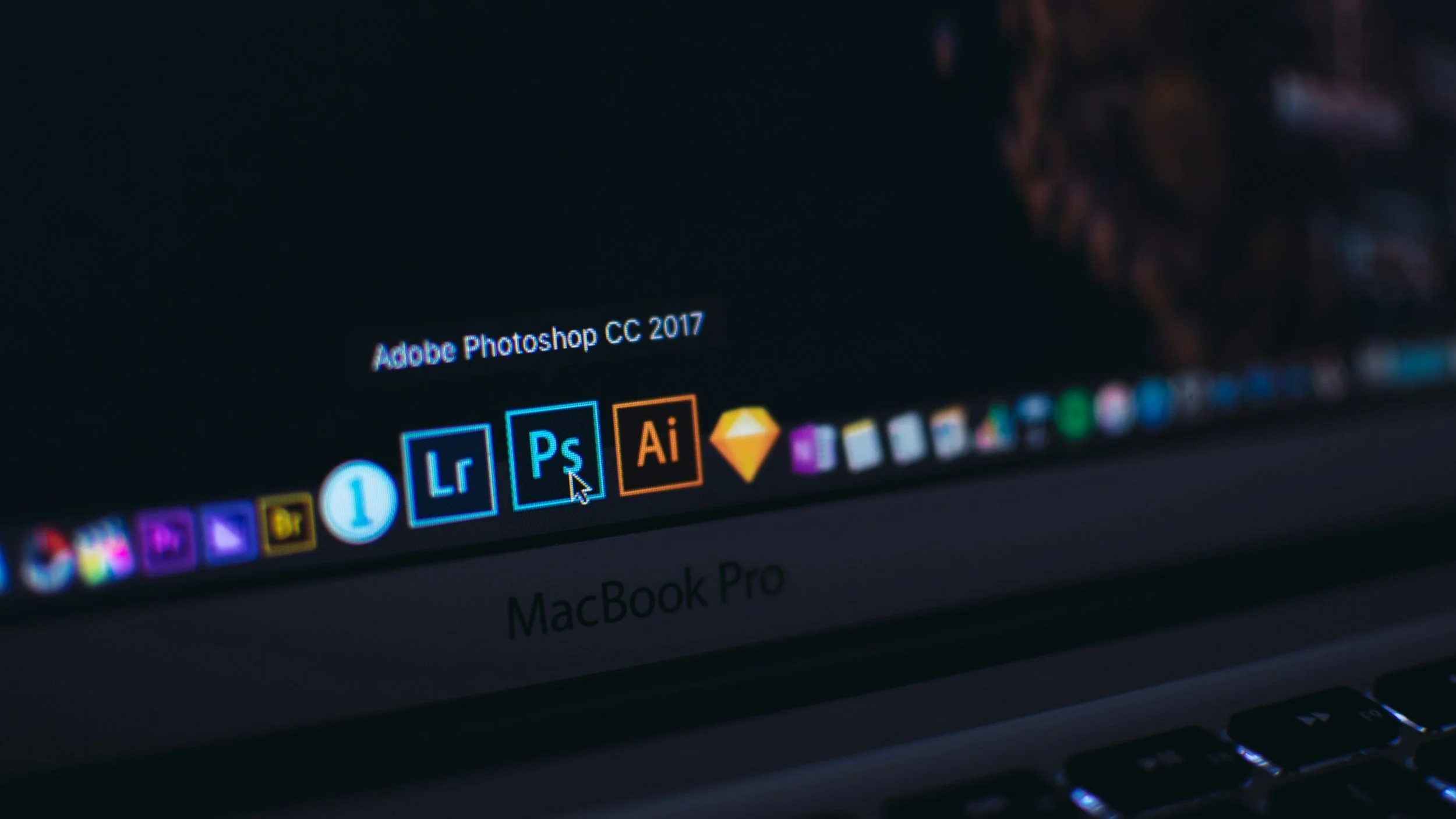 Close-up of a MacBook Pro screen displaying Adobe Photoshop CC 2017, with application icons including Lightroom, Illustrator, and Sketch visible on the dock.
