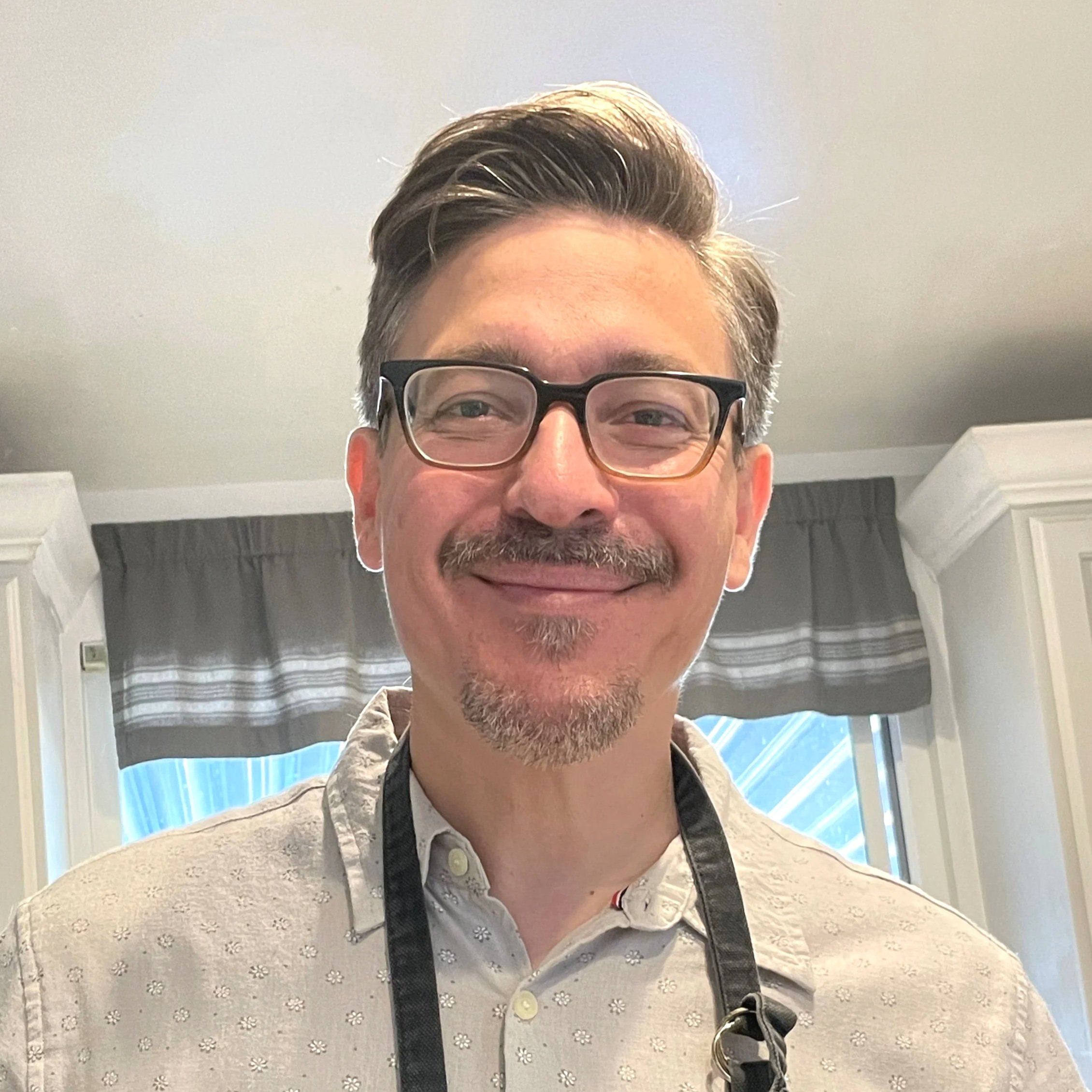 A man with glasses and a goatee smiling in front of a window with curtains.