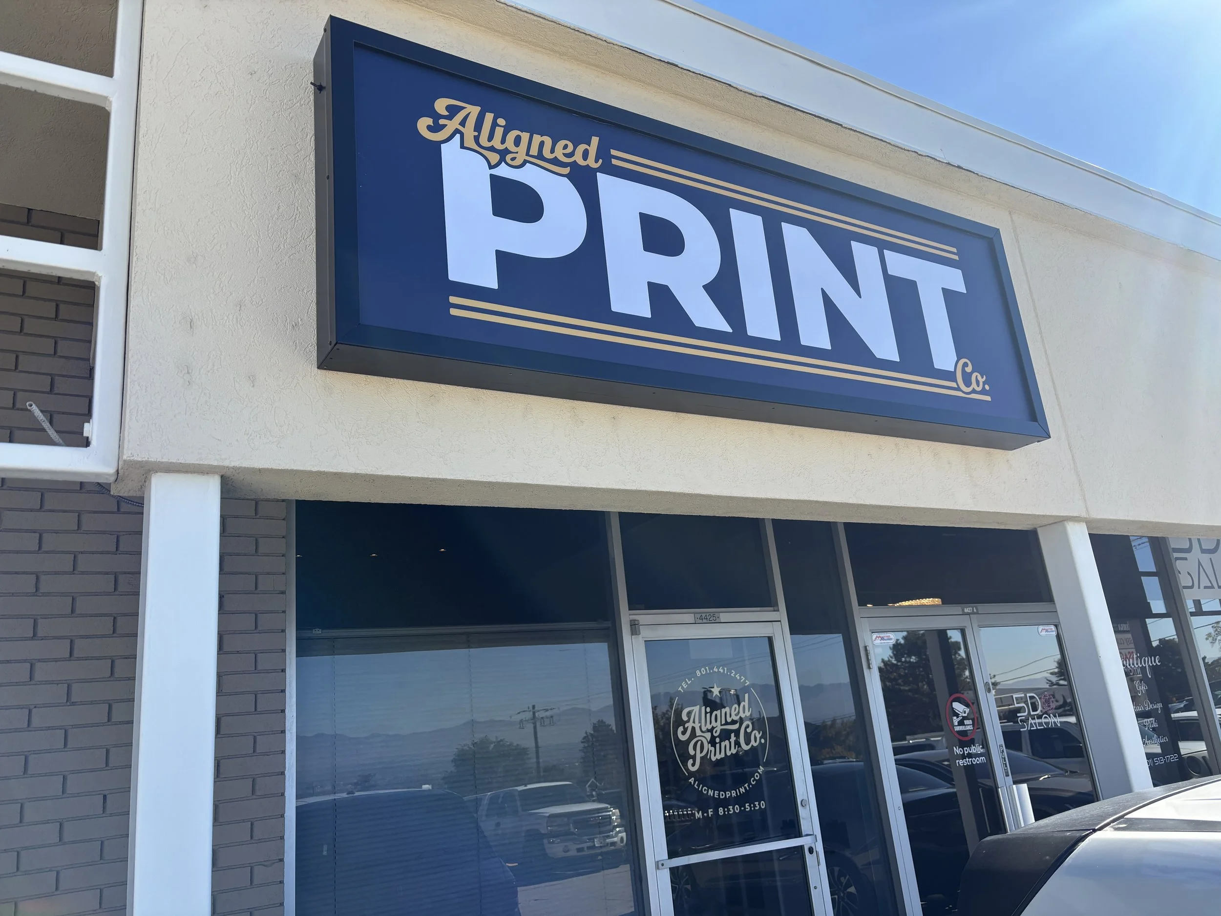 Exterior of a copy and print shop with a large sign that reads 'Aligned PRINT Co.' above the entrance. The shop has glass doors and windows, and a reflection of parked cars in Salt Lake City and a mountain range can be seen in the glass.