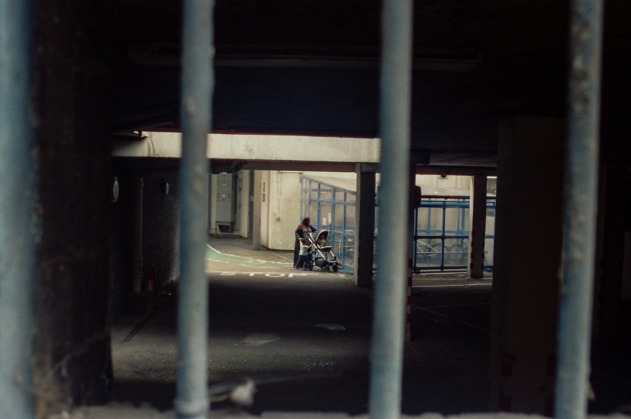 A person standing with a stroller near a bicycle rack, seen through metal bars under an overpass or parking garage