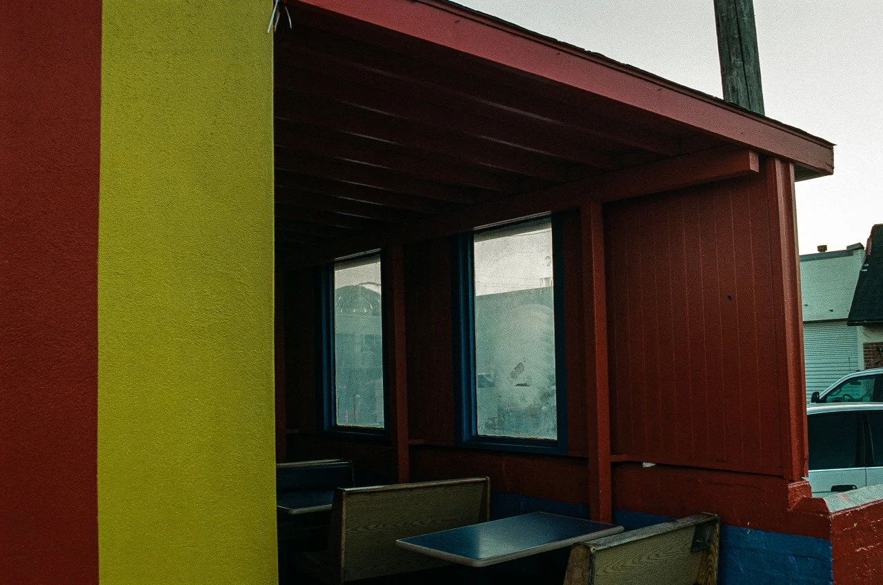 Interior of a small restaurant or cafe with red and yellow painted walls, featuring a window and seating area with chairs and tables.