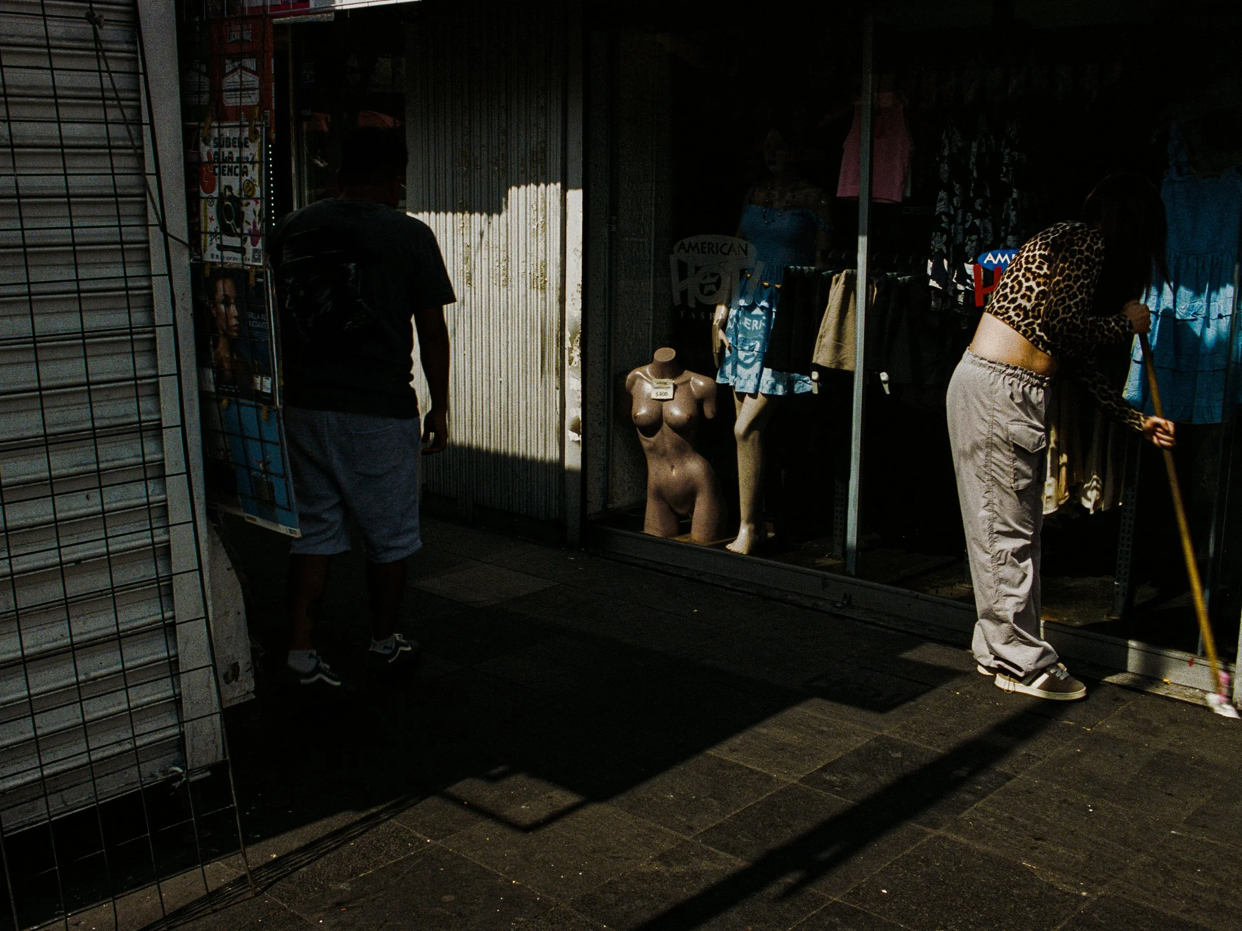 A clothing store display with mannequins and garments on racks, seen through a storefront window with reflections and shadows.