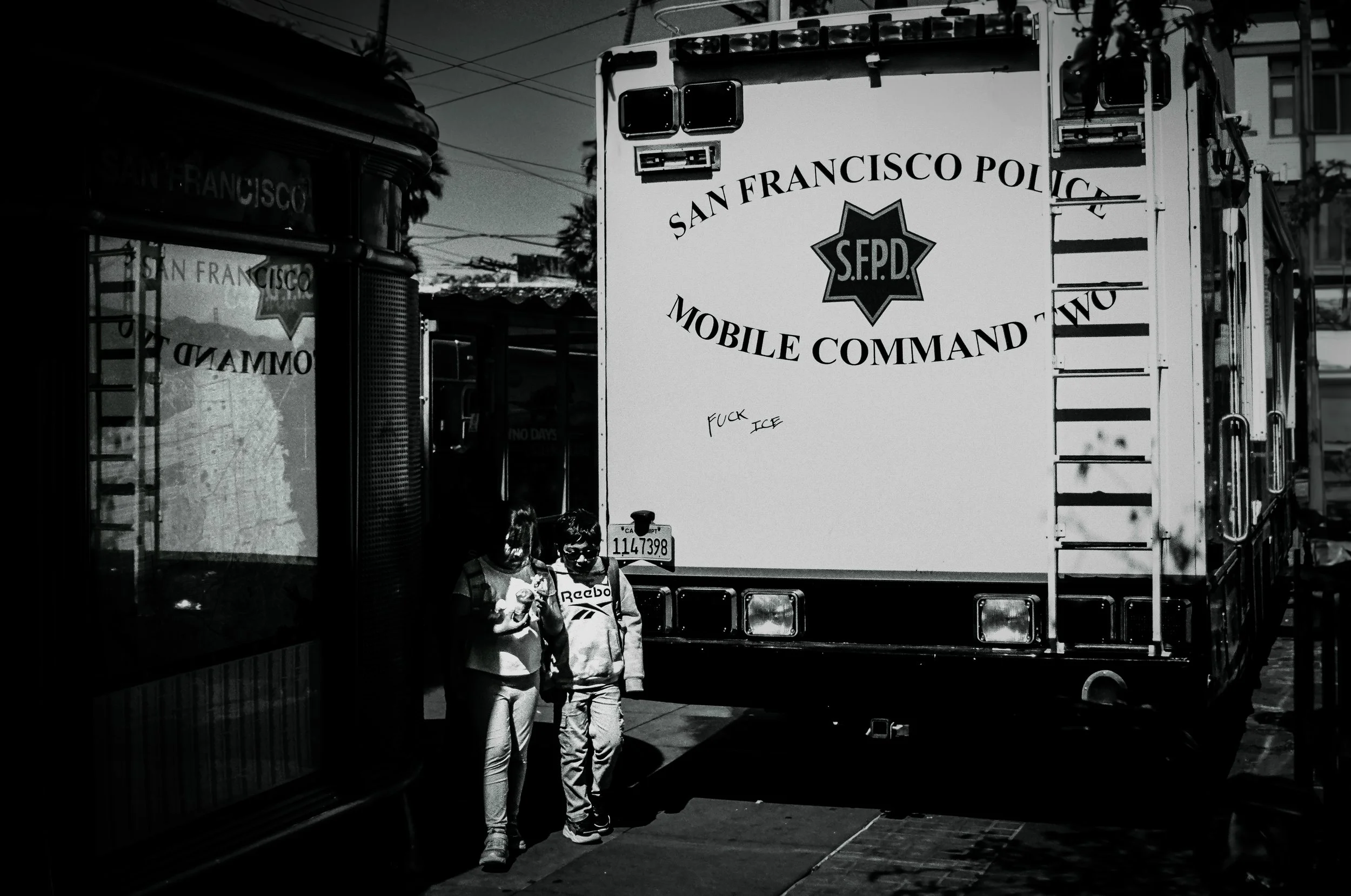 Black and white photo of San Francisco police mobile command vehicle with two children standing in front of it, wearing casual clothing and sunglasses, with one child holding a drink. The police vehicle has the words 'Santa Francisco Police', 'S.F.P.