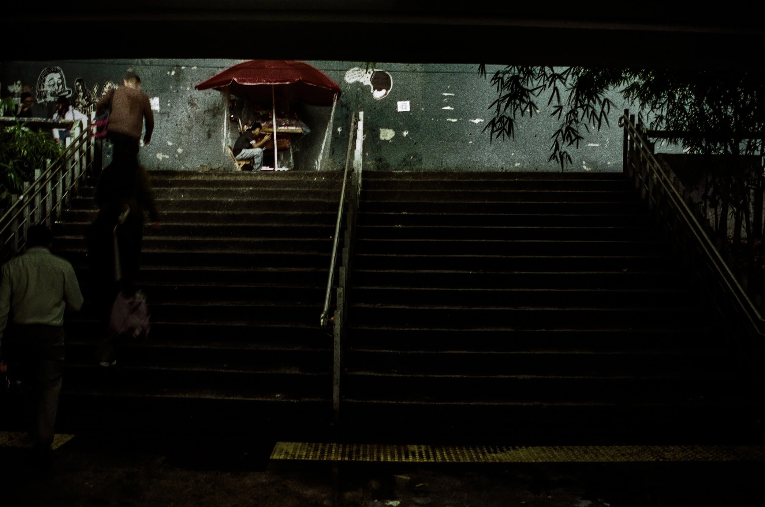Nighttime scene under an overpass featuring a set of dark stairs leading up to a graffiti-covered wall. A person with a backpack and several others are walking or standing around, with some seated at a small food stall with a red umbrella. The overal
