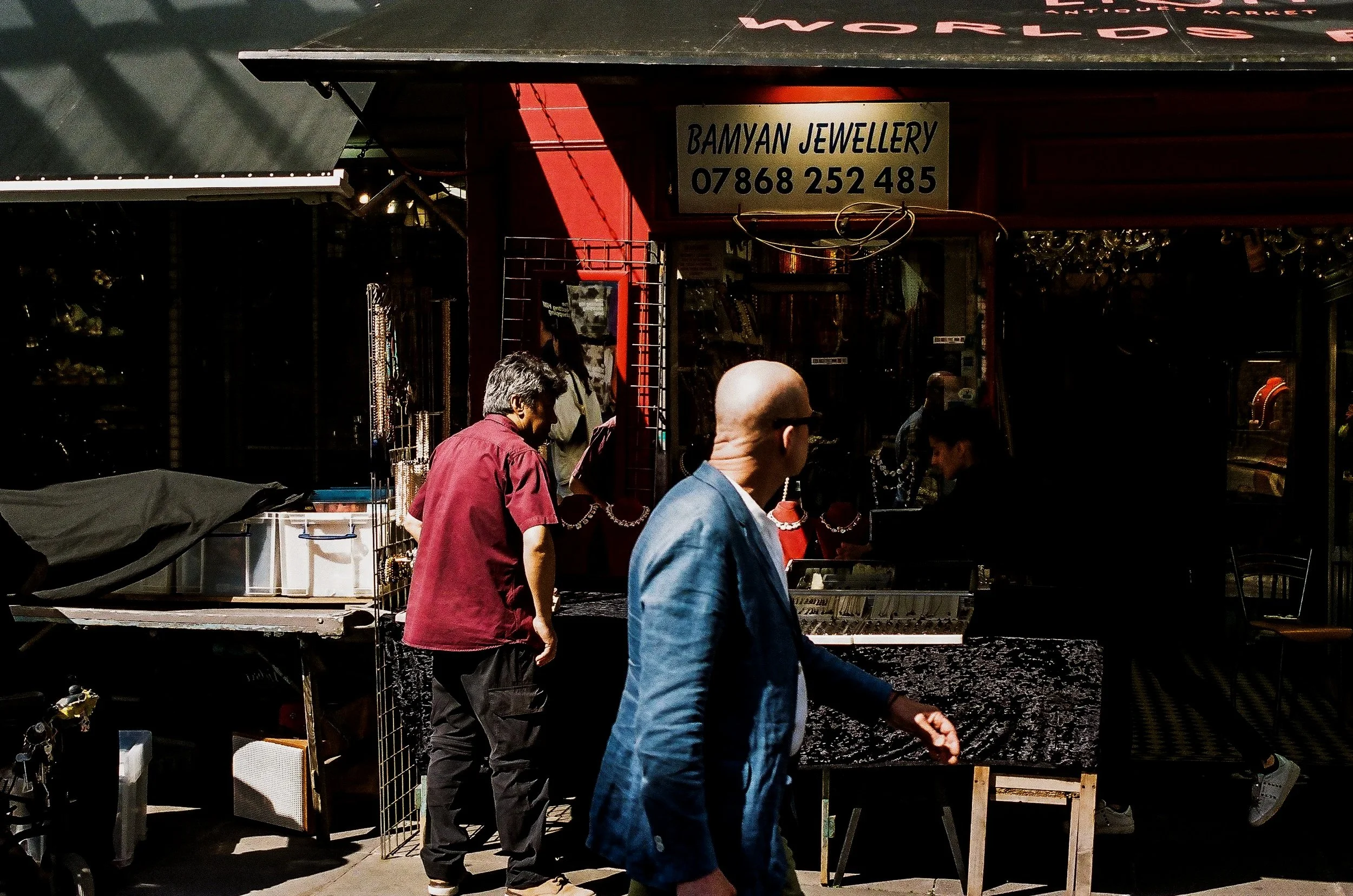 People shopping at a jewelry stall labeled 'Bamyan Jewelry' with a phone number, displaying various jewelry pieces.