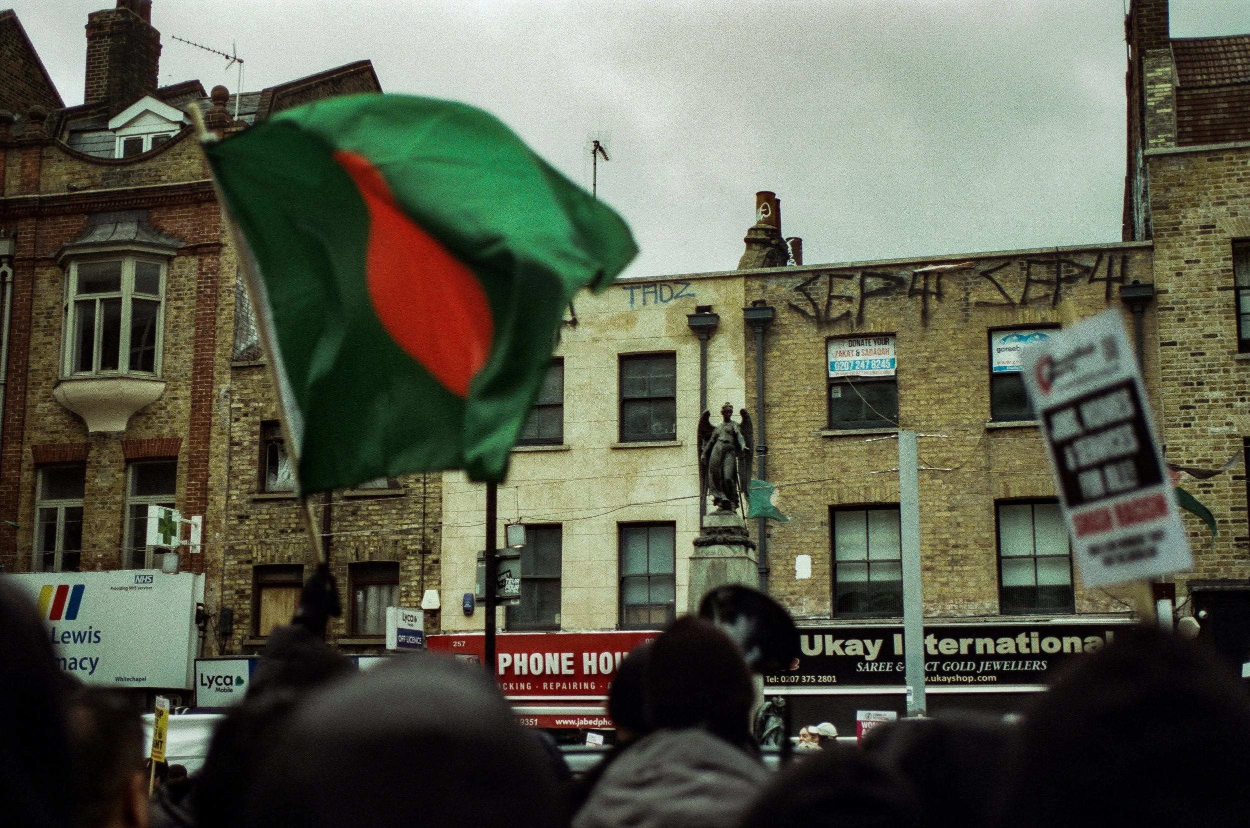 A crowd of people gathered in a city square with buildings in the background, some graffiti on the walls, and a green umbrella with a red stripe in the foreground.