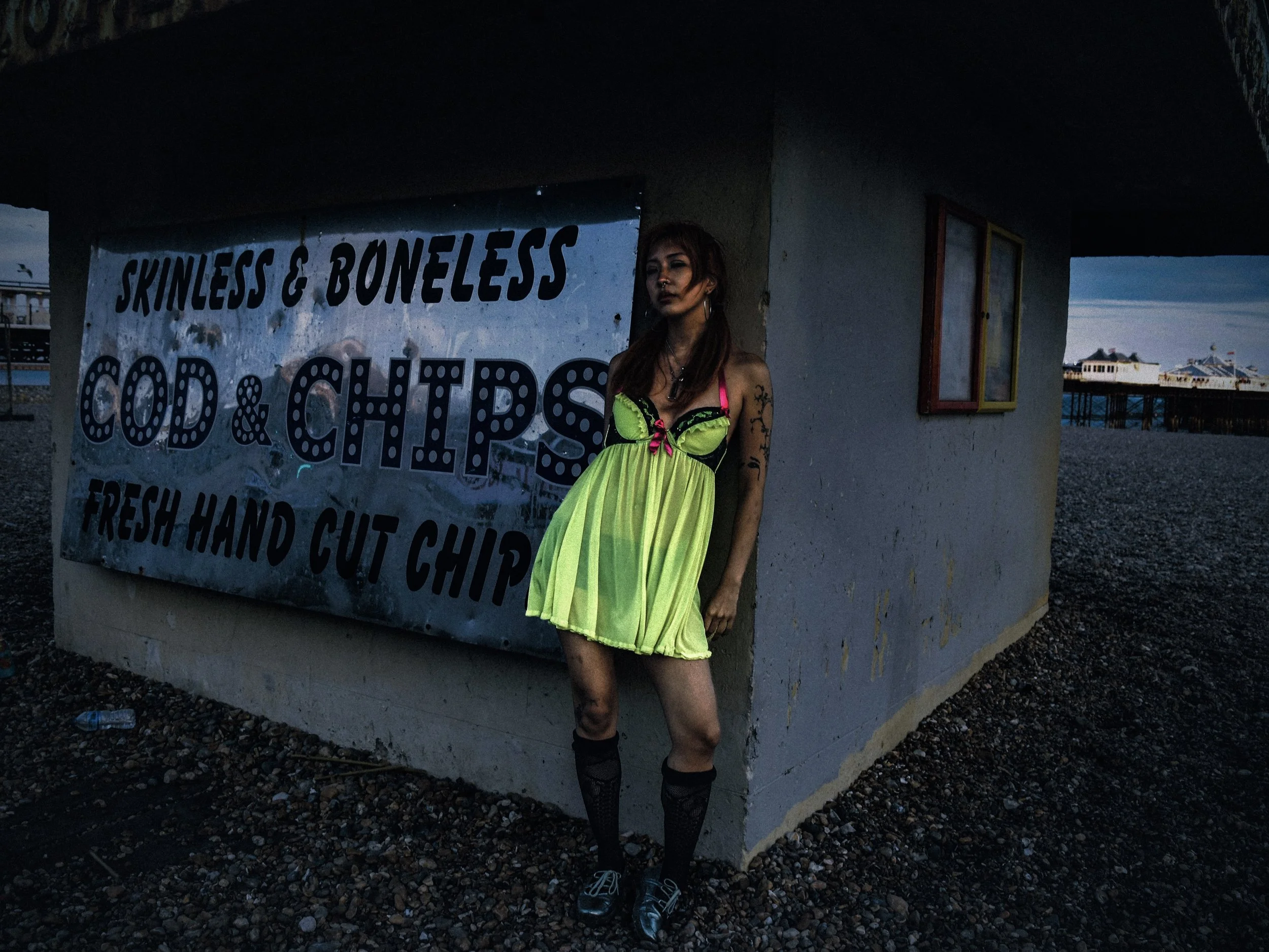 A young woman with tattoos and dark makeup leaning against a sign at an outdoor location during sunset. The sign advertises "Skinnless & Boneless Cold & Chips" and mentions fresh hand-cut chips." in a coastal or pier area.