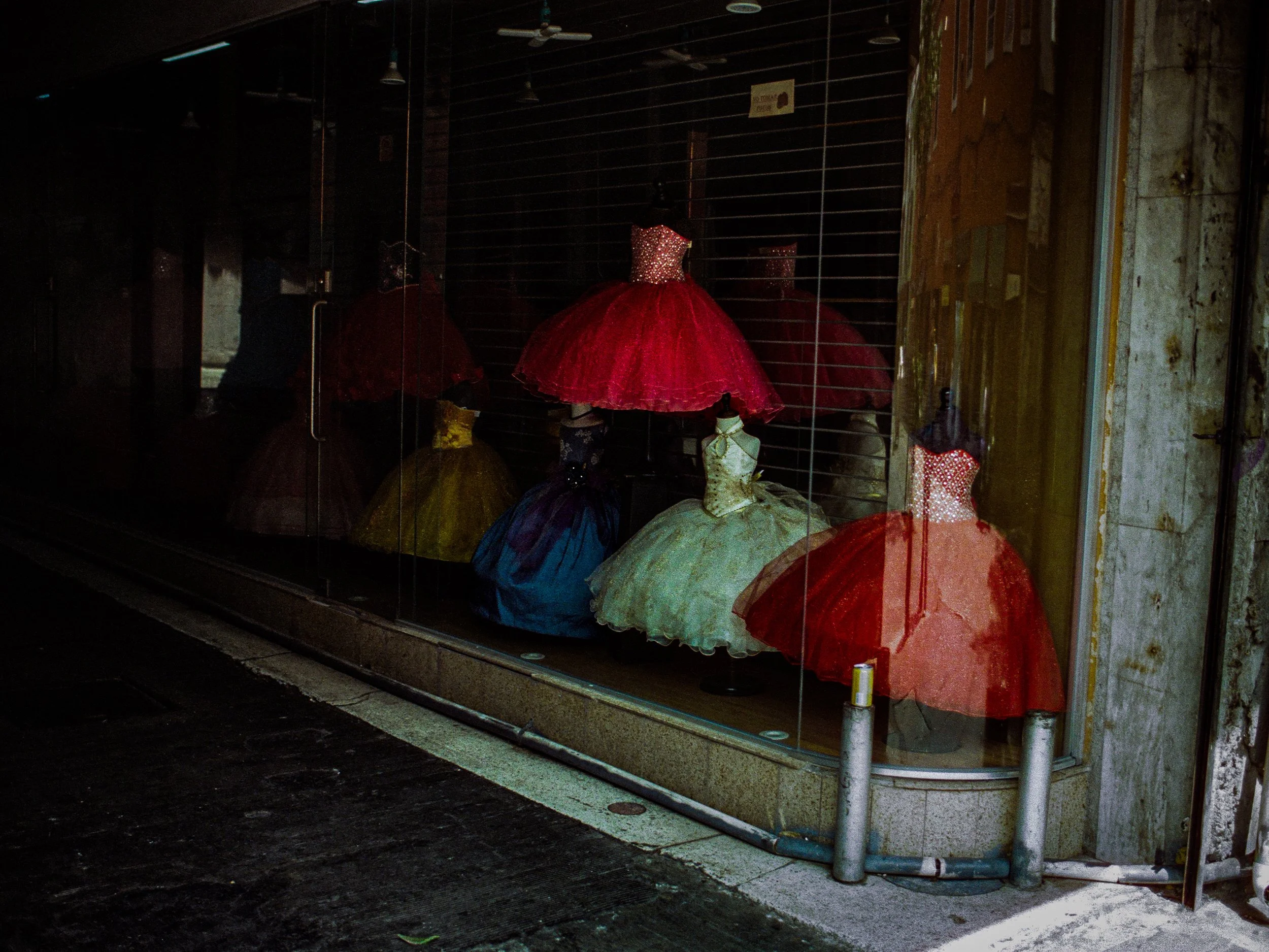 Display window with several colorful dresses on mannequins, including a prominent red dress with a sparkly bodice. The window has a reflective glass surface showing nearby street and buildings.