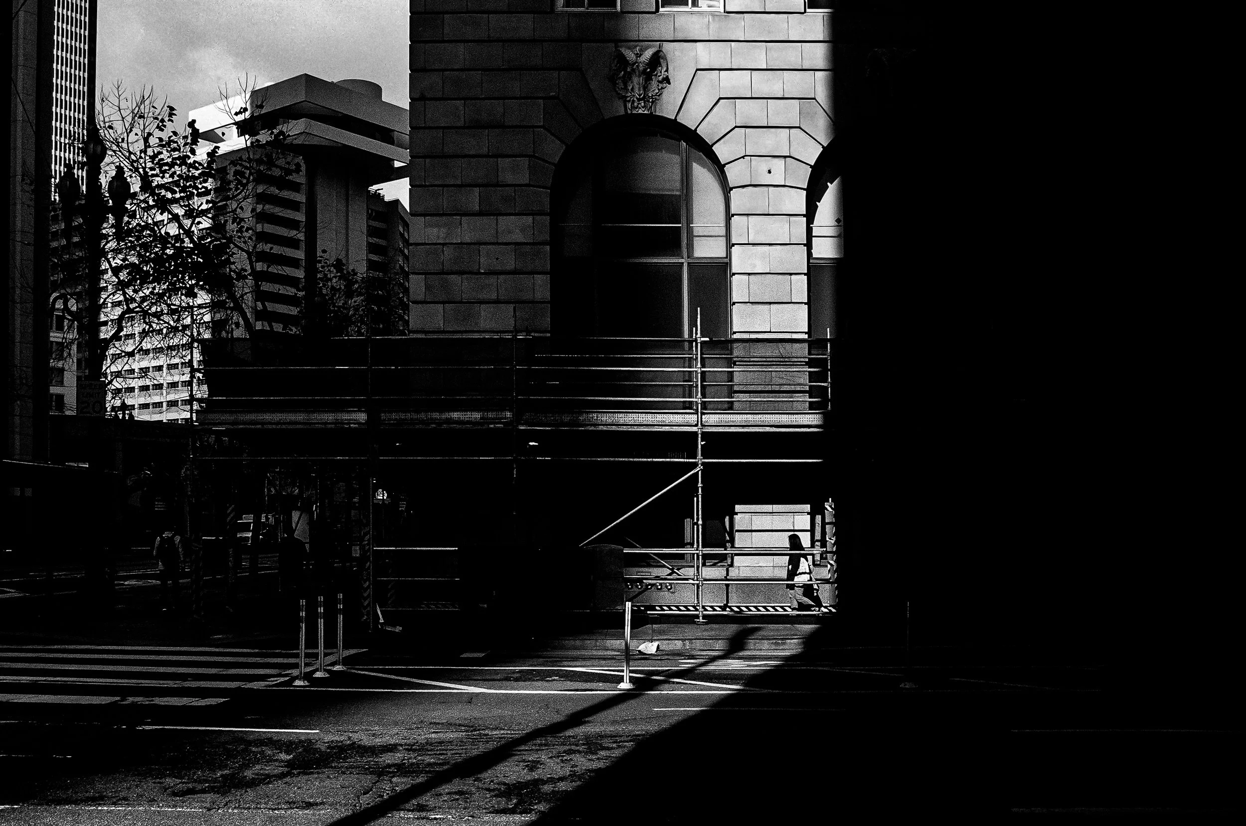 Black and white photo of an urban scene with buildings, trees, and a person walking on the sidewalk. Scaffolding is present in the foreground, partially blocking the view of a large window and stone facade of a building.