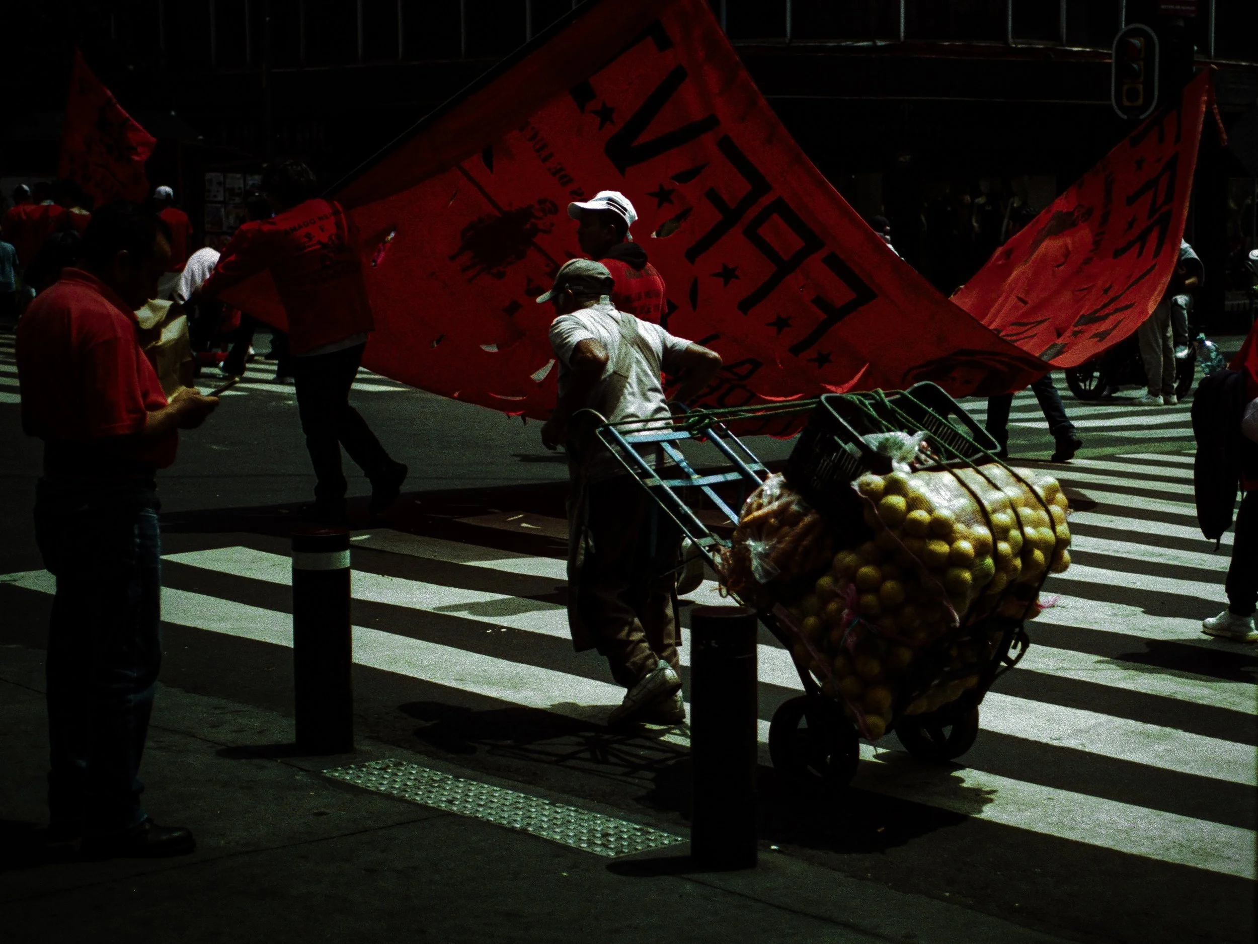 People crossing a city street at a crosswalk, with a person pushing a shopping cart filled with produce and a large red flag in the background.
