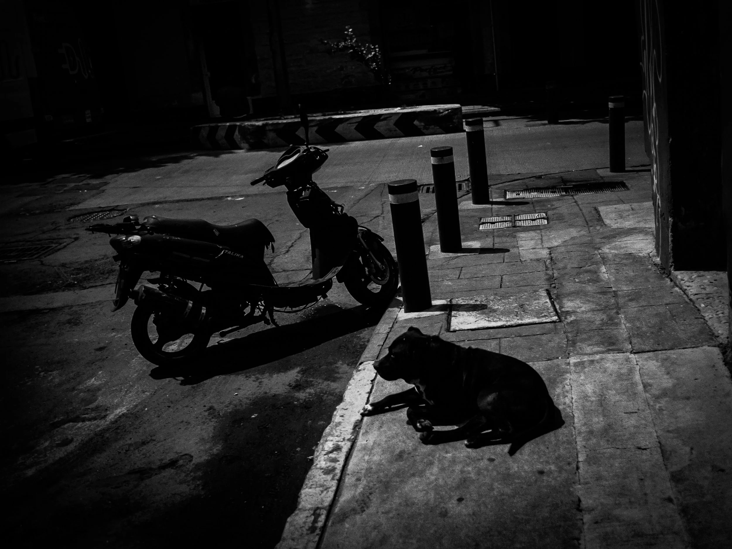 A black and white photo of a motorcycle parked on a city sidewalk at night, next to a curb and several bollards, with a building and fence in the background.