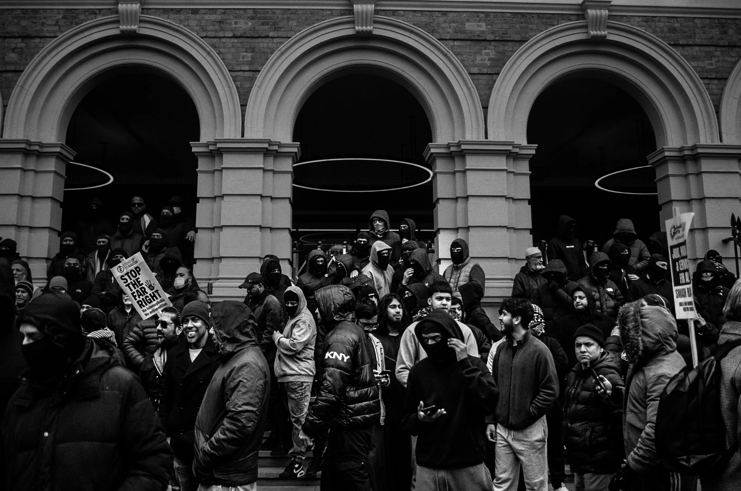 A large group of protesters gathered in front of a building with arched windows, many of whom are wearing black masks and holding signs, including one that reads 'Stop the Far Right'.