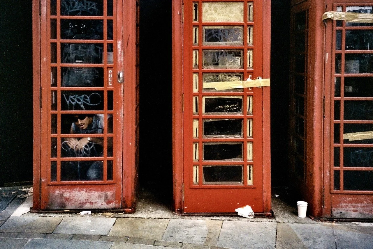 A row of red telephone booths, with one booth’s door slightly open and a person inside wearing sunglasses, crouching and looking out.