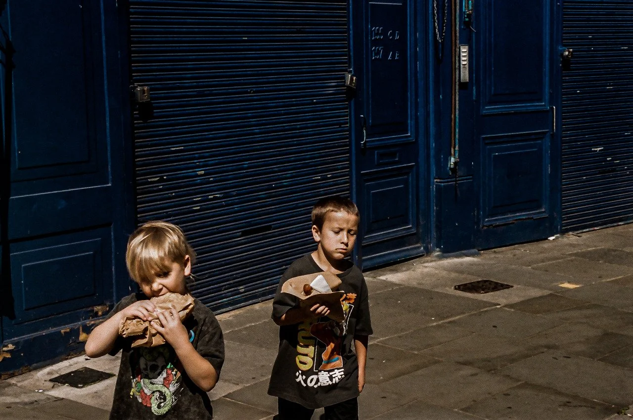 Two young boys standing on a sidewalk in front of dark blue storefronts with closed shutters. One boy is holding a wrapped sandwich, appearing to eat or about to eat, while the other boy is holding a paper bag and looks pensive or sad.